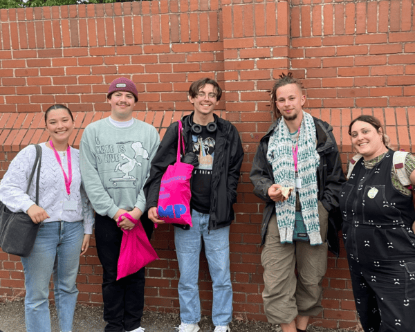 The picture shows four participants from the AMP Producers programme standing against a brick wall outside the music venue they completed their event at. On the right there is also Teah (a 30 year old woman with dark hair, wearing a jumpsuit and has a backpack on). She is the AMP Programme manager who worked with the young people on this event. The young people are smiling, they are wearing AMP lanyards and have AMP tote bags which are bright pink with electric blue text that says AMP. 